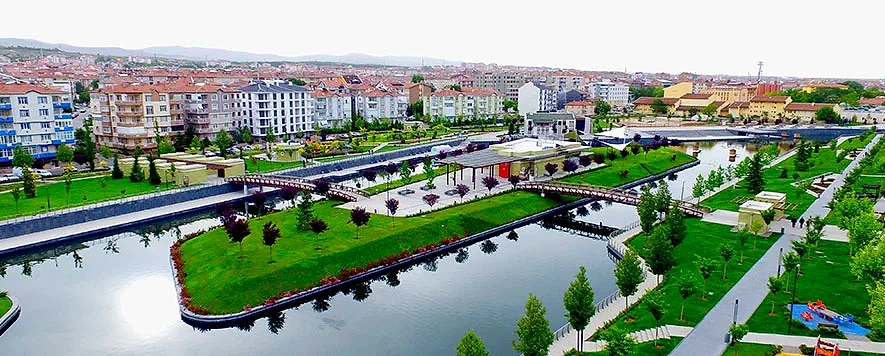 Şehir parkı ve çevresi hakkında bilgi Şehir parkı manzarası, yeşil alanlar ve su kenarıyla çevrili modern yapılar.